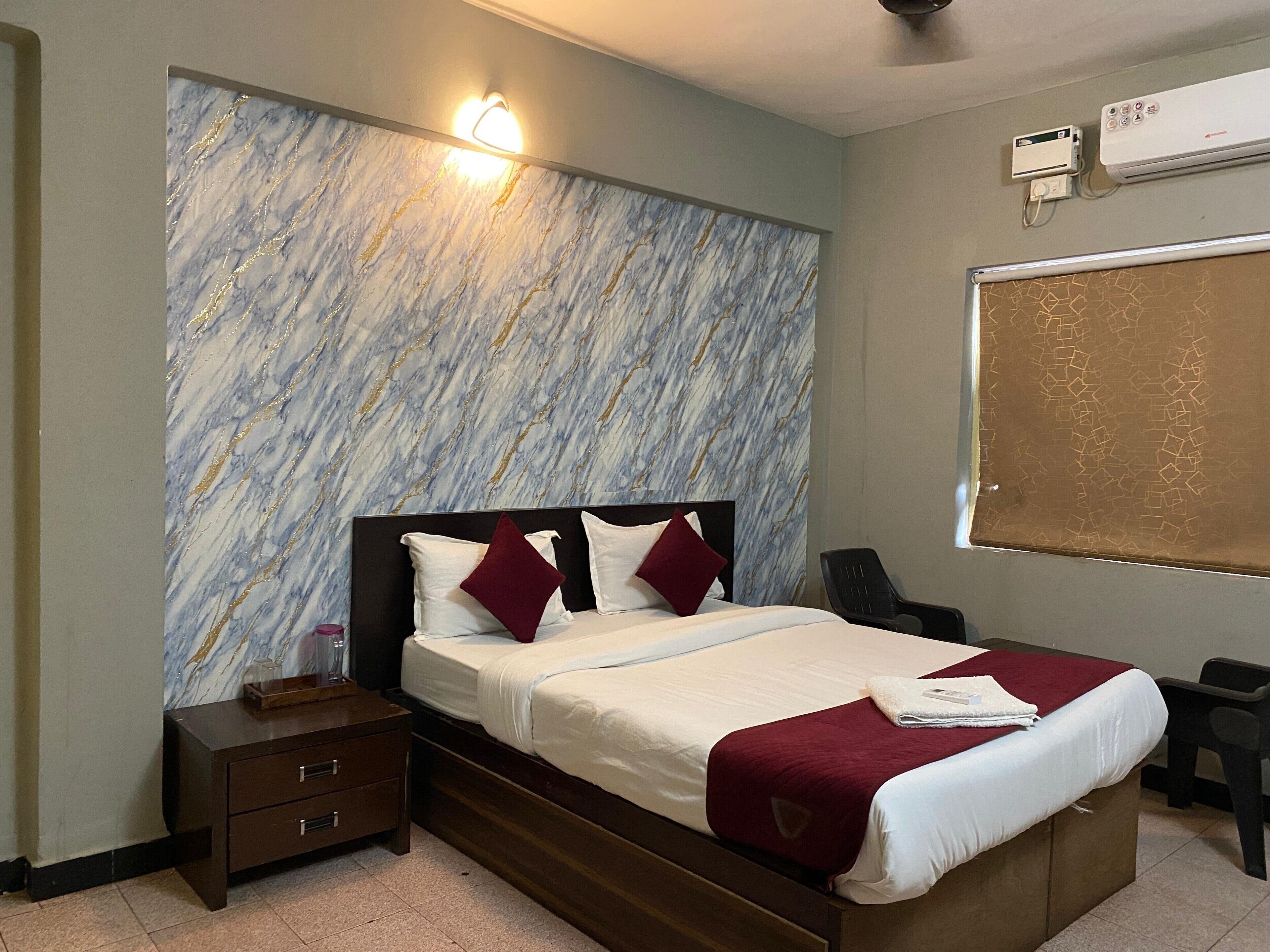 A hotel room with a marble-patterned wall, a bed with white bedding and red accents, a wooden nightstand, and a desk with a chair.