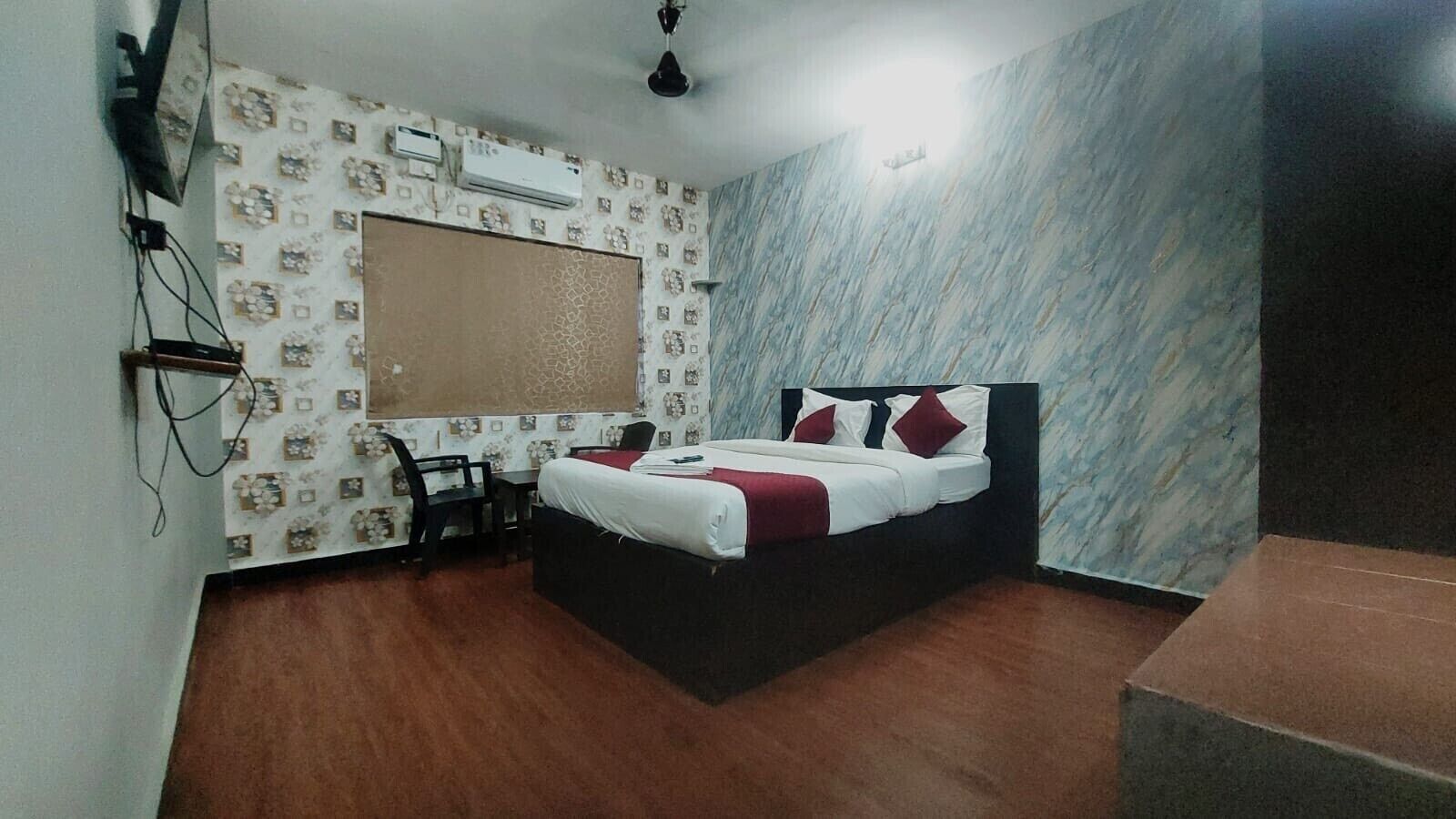 A bedroom with a bed, a desk, and a chair. There is a window with a curtain and an air conditioning unit on the wall.