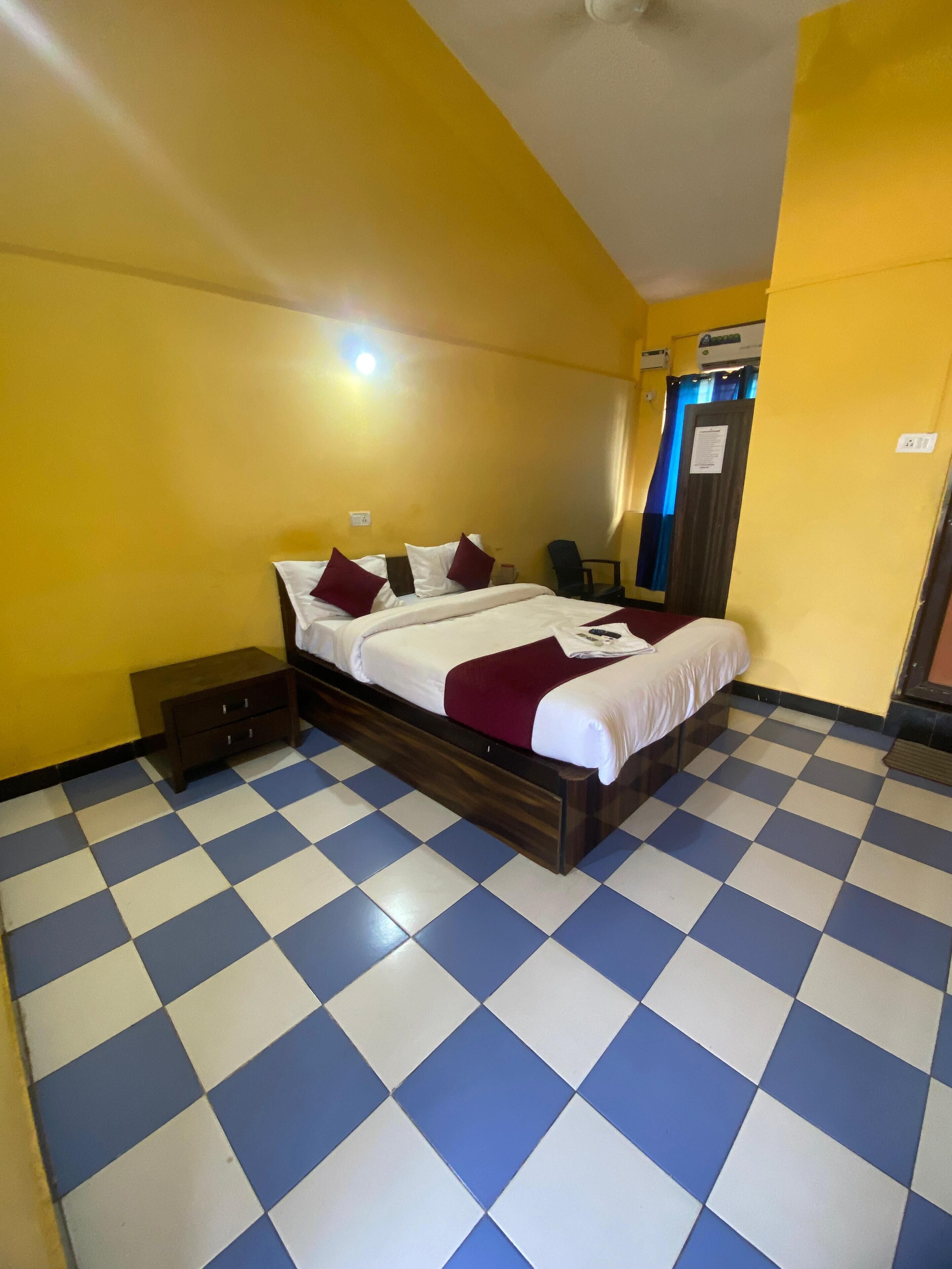 A hotel room with a checkered floor, a bed with white and maroon bedding, a nightstand, a chair, and a door with a sign.