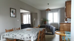 Et spiseområde med et bord dækket af en blomstret dug, træstole, en sofa samt et køkken med træskabe og en vask.