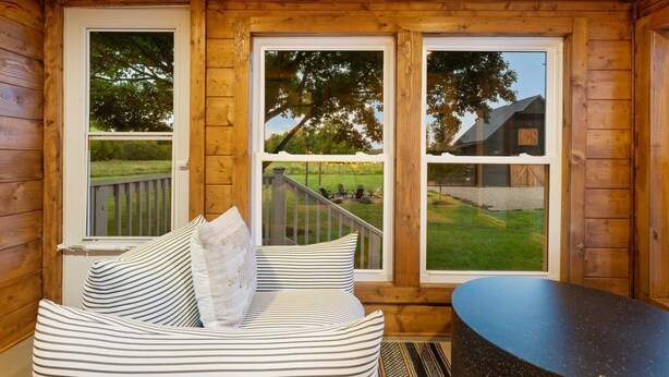Una cabaña de madera con sofá y mesa, ventanas amplias con vista a un patio cubierto de césped y un edificio.