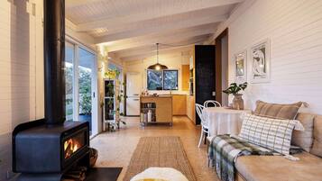 A cozy living room with a fireplace, a sofa, a coffee table, and a kitchen area with a refrigerator and a hanging light fixture.