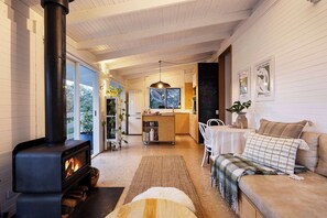 A cozy living room with a fireplace, a sofa, a coffee table, and a kitchen area with a refrigerator and a hanging light fixture.