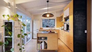 A modern kitchen with a large island, a blackboard with 'Welcome' written on it, and a view of the outdoors through large windows.