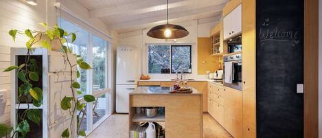 A modern kitchen with a large island, a blackboard with 'Welcome' written on it, and a view of the outdoors through large windows.