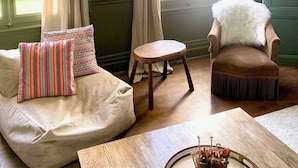 Una habitación con una puerta verde, un sofá beige con cojines estampados, una silla de madera con una manta de piel blanca y una pequeña mesa de madera con una bandeja decorativa.