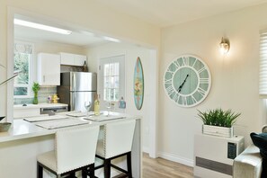 A modern kitchen with white cabinetry, a stainless steel refrigerator, and a dining area with a white table and chairs. A large wall clock and a surfboard decoration are visible.