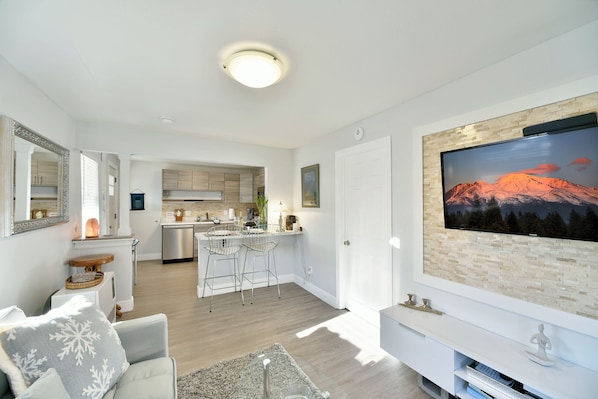 A modern living room with a large flat-screen TV mounted on a brick wall, a cozy seating area with a sofa and a coffee table, and a kitchen area with wooden cabinets and a dishwasher.