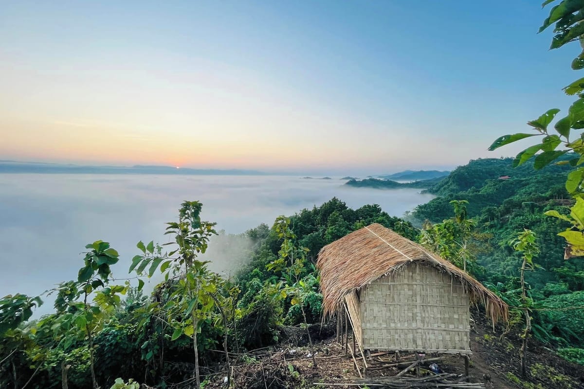 A thatched-roof hut with a view of a mist-covered landscape and distant hills.