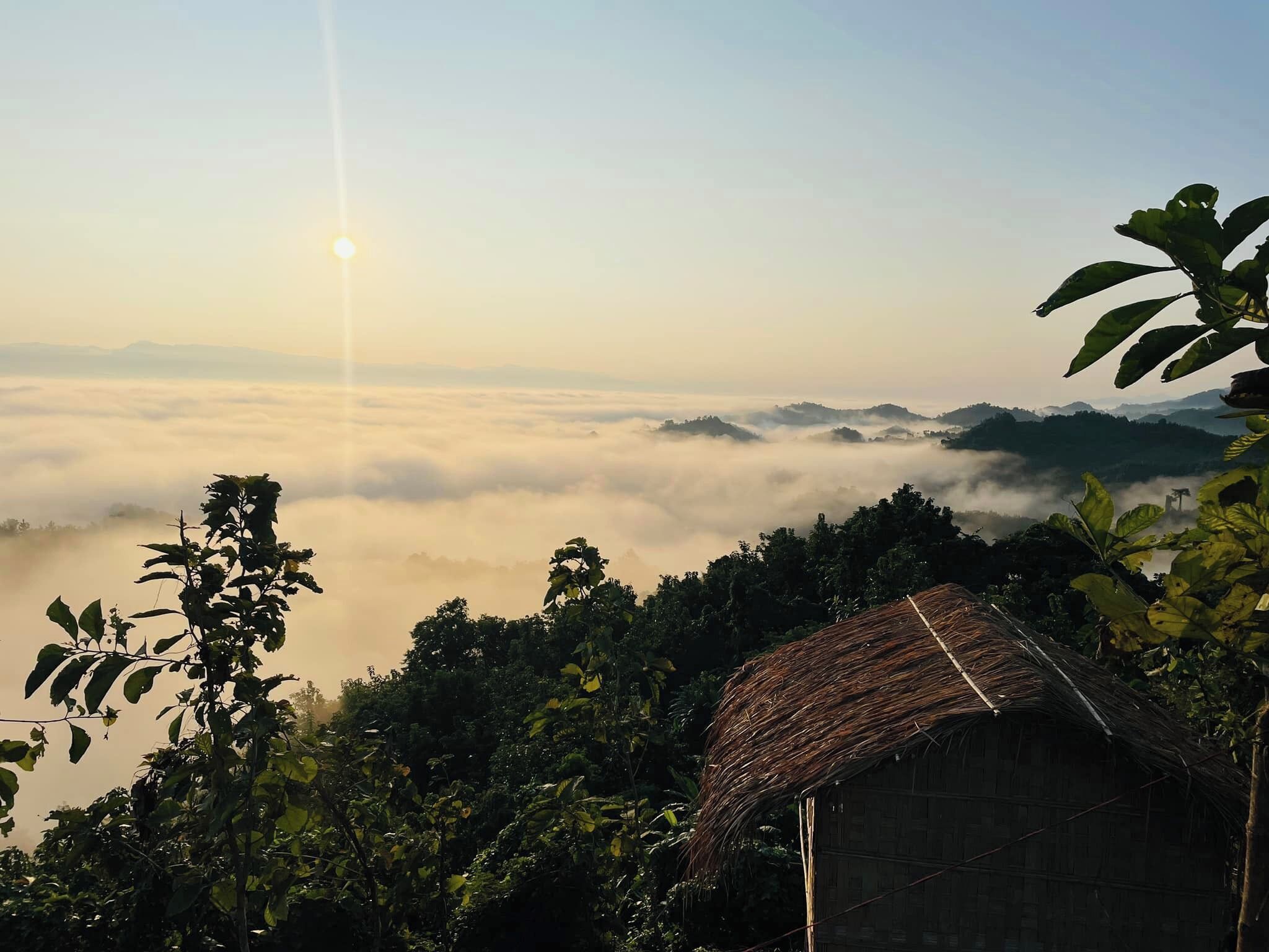 Uma cabana com telhado de palha, rodeada por vegetação densa e uma paisagem montanhosa envolta em névoa.