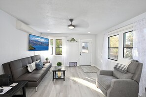 Un salon moderne avec un canapé, un fauteuil et une table basse. Un téléviseur est fixé au mur et une porte donne sur l’extérieur.