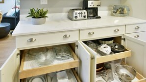 Une cuisine avec des meubles blancs, un plan de travail sur lequel on trouve un grille-pain, une machine à café et une plante. On y voit plusieurs étagères remplies de verres et une pancarte indiquant « Café ».