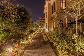 A nighttime view of a pathway flanked by lush greenery and a building with multiple windows and balconies.