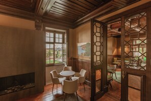 A room with a fireplace, a round table with chairs, and a large window with a view of greenery.