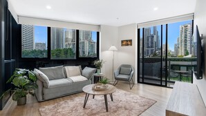 A modern living room with a grey sofa, a small round table, and a floor lamp. Large windows offer a view of tall buildings and greenery.