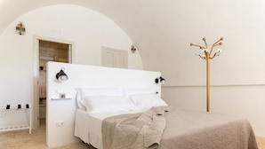 Ein Schlafzimmer mit einem weißen Bett, einer beigen gesteppten Tagesdecke, einem hölzernen Kleiderständer und einer weißen Tür.