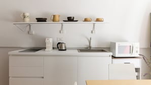 Eine moderne Küche mit weißen Schränken, einer weißen Arbeitsplatte, einem Wasserkocher, einer Kaffeemaschine, einer Mikrowelle und einem Regal mit verschiedenen Tassen und Schüsseln.