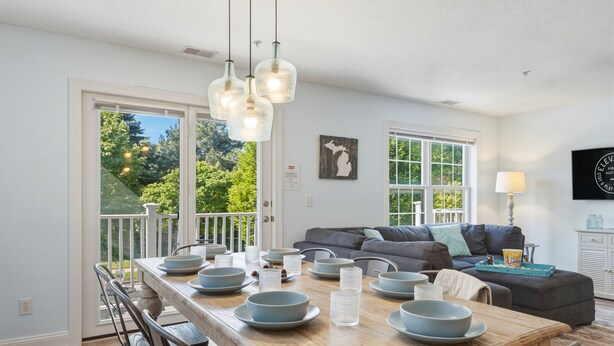Un coin repas avec une table en bois, dressée avec des bols et des assiettes, un canapé gris et une vue sur la verdure à l’extérieur.