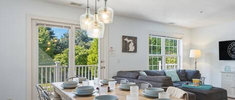 A dining area with a wooden table set with bowls and plates, a gray sofa, and a view of greenery outside.