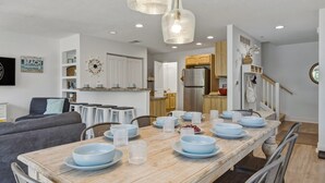 Un coin repas avec une table en bois dressée pour six personnes, entourée de chaises, et une cuisine en arrière-plan.