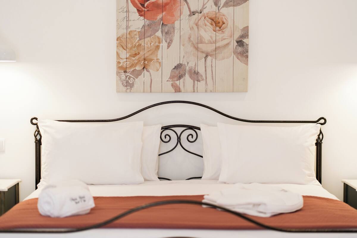 A neatly made bed with white linens and a brown bedspread, a floral artwork on the wall, and a black metal headboard.
