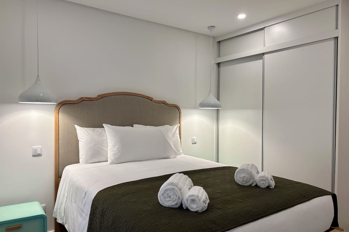 A neatly made bed with white linens, a dark green blanket, and rolled towels. There is a teal bedside table with a drawer, a wall-mounted light, and a ceiling light.