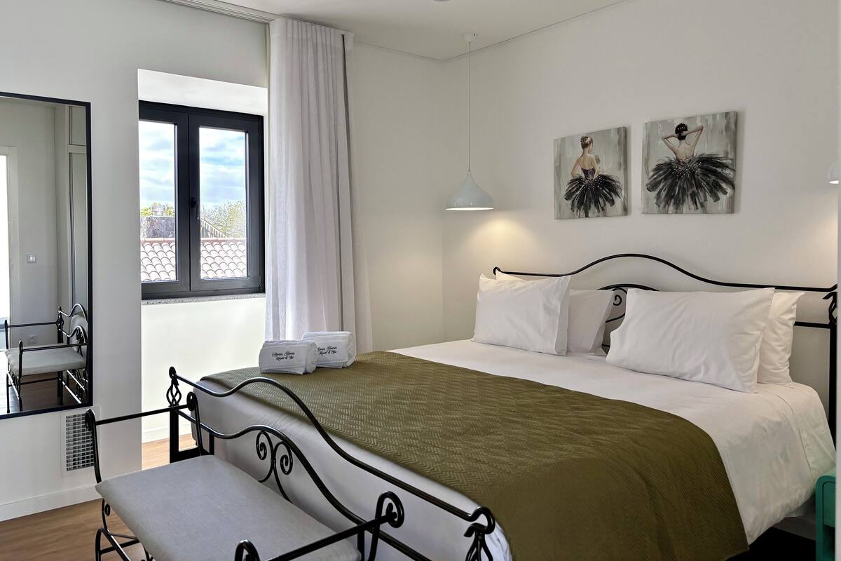 A neatly arranged bedroom with a bed, a bench, a window with curtains, and artwork on the wall.