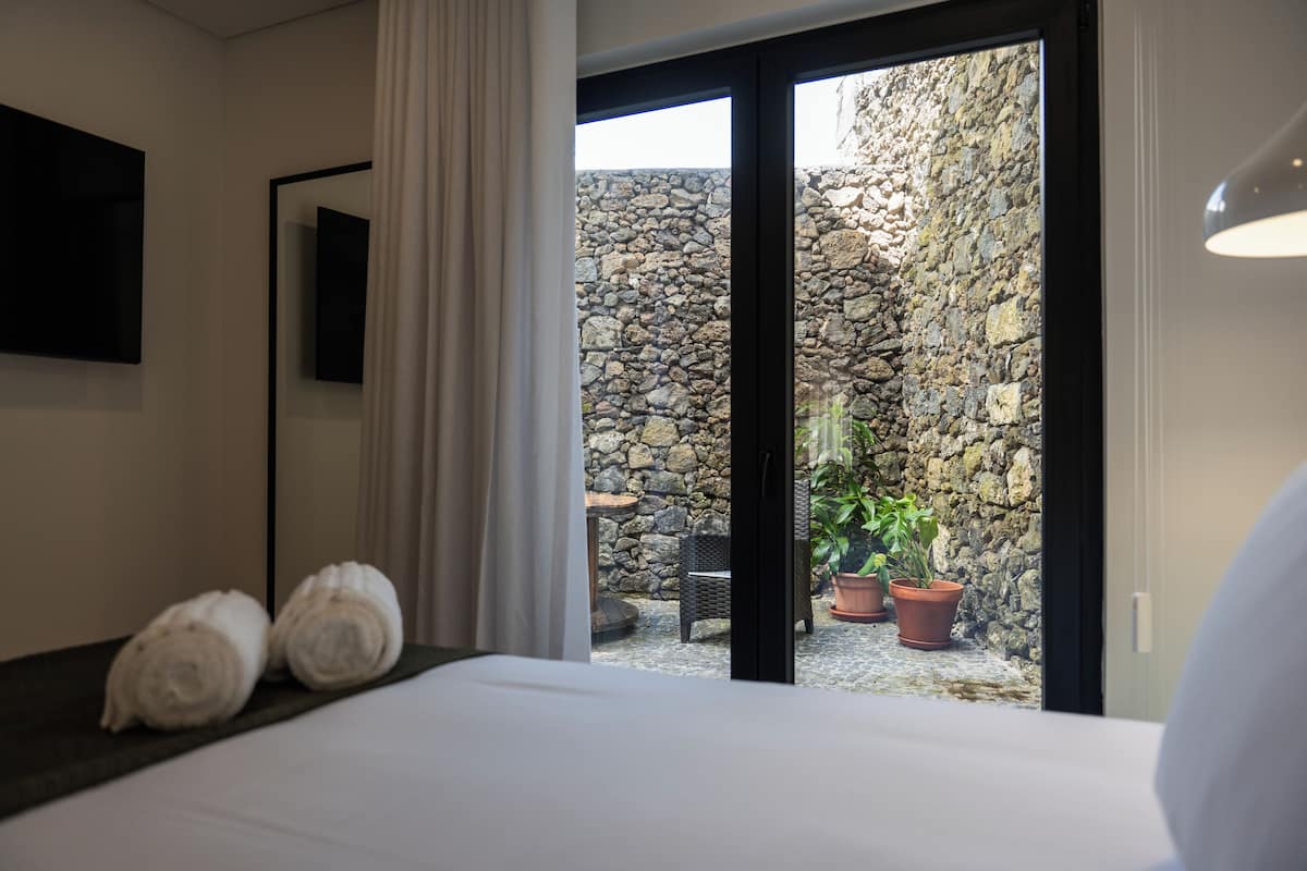 A hotel room with a bed, a flat-screen TV mounted on the wall, and a view of a stone wall and potted plants outside.