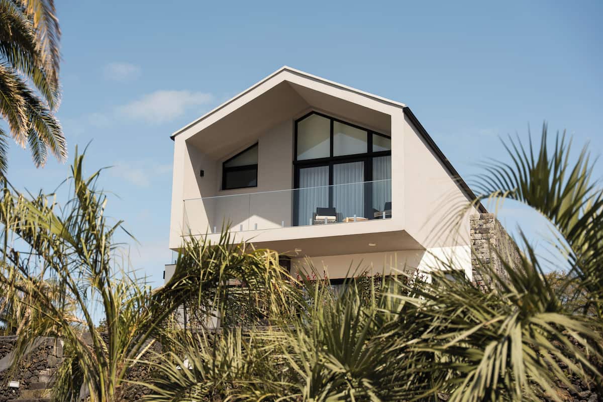 A modern two-story house with a balcony, surrounded by tropical vegetation.