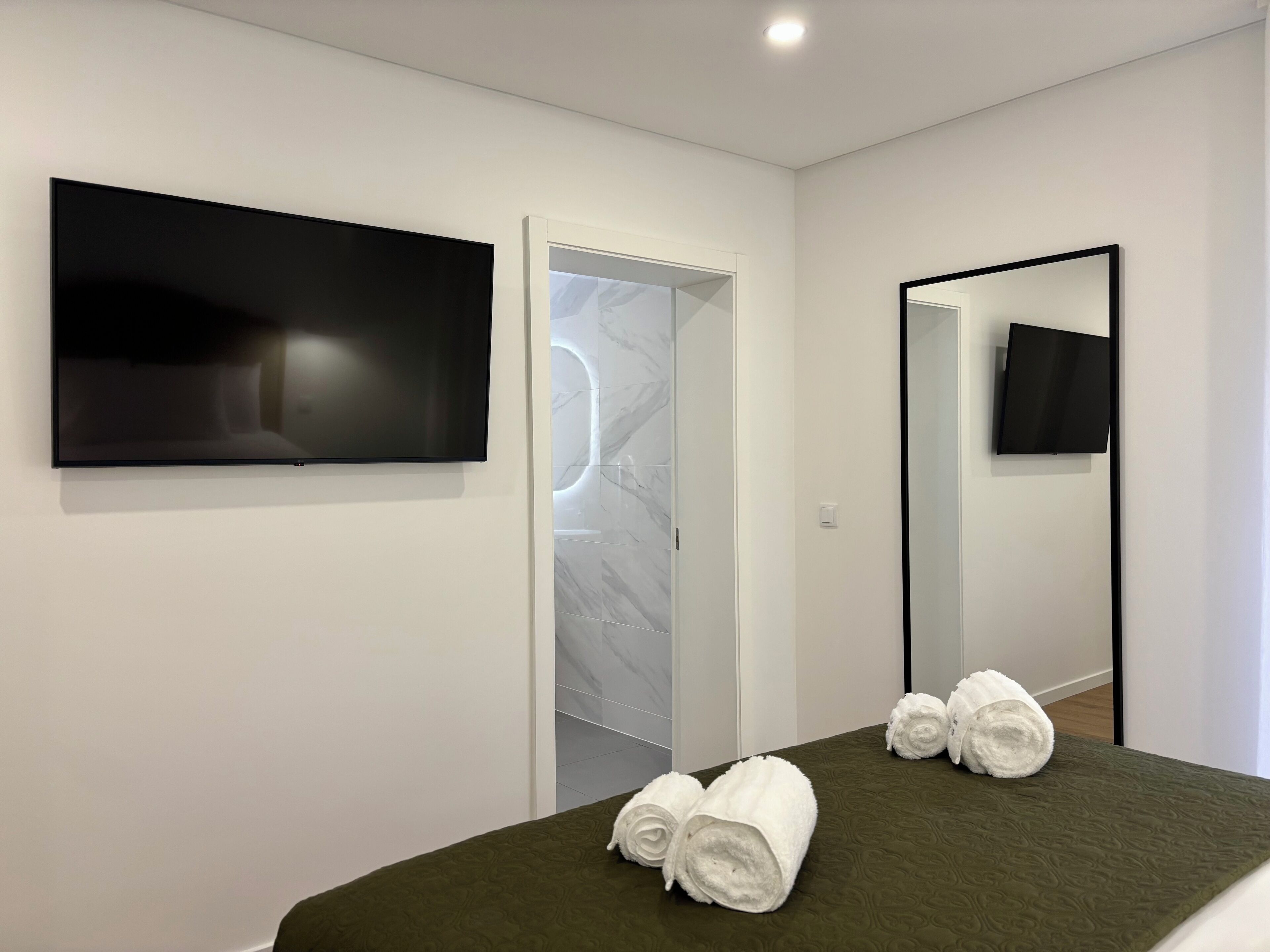 A modern hotel room with a bed, a flat-screen TV mounted on the wall, a mirror, and a door leading to a bathroom.