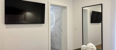 A modern hotel room with a bed, a flat-screen TV mounted on the wall, a mirror, and a door leading to a bathroom.