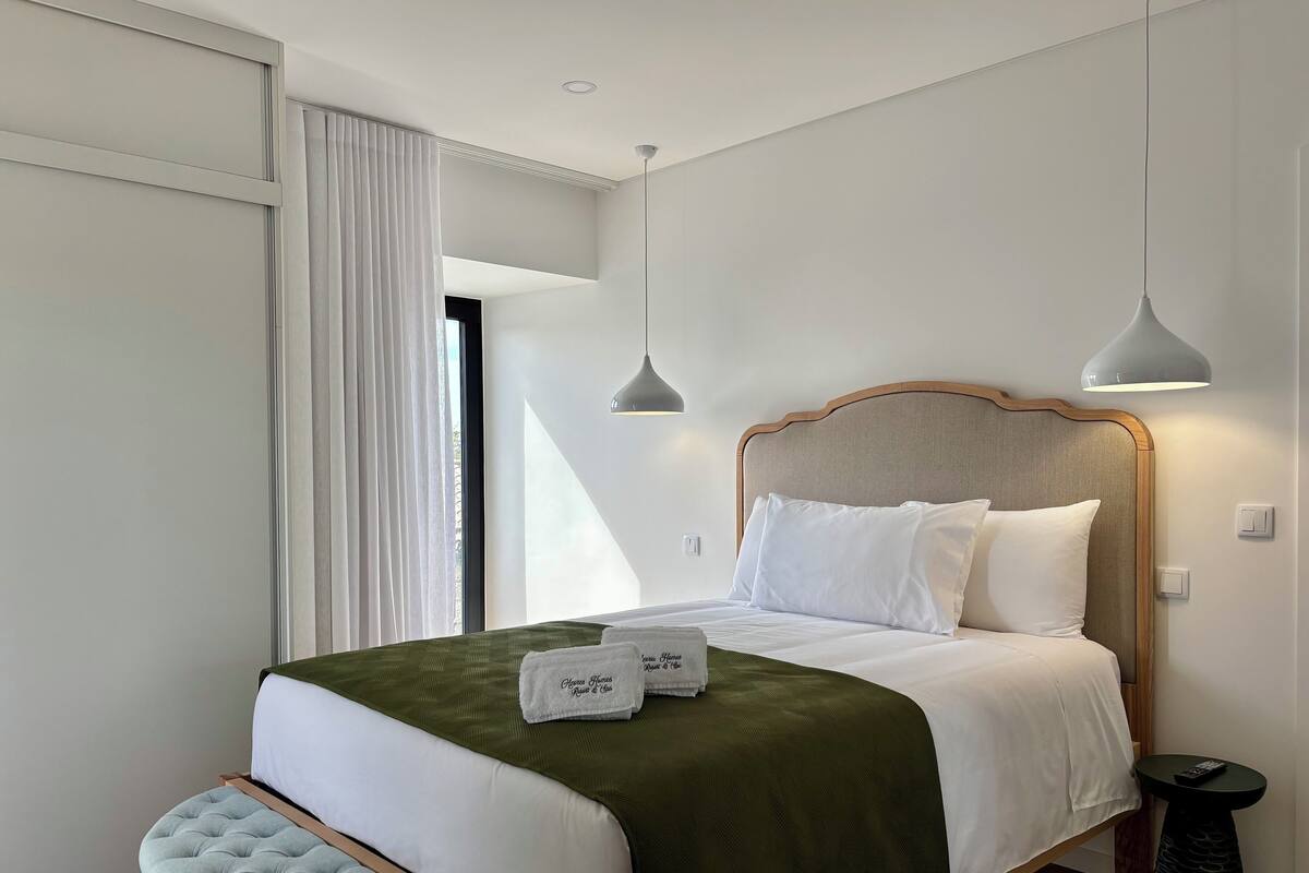 A neatly made bed with a green blanket and white pillows, a bedside table with a remote control, and a window with white curtains.