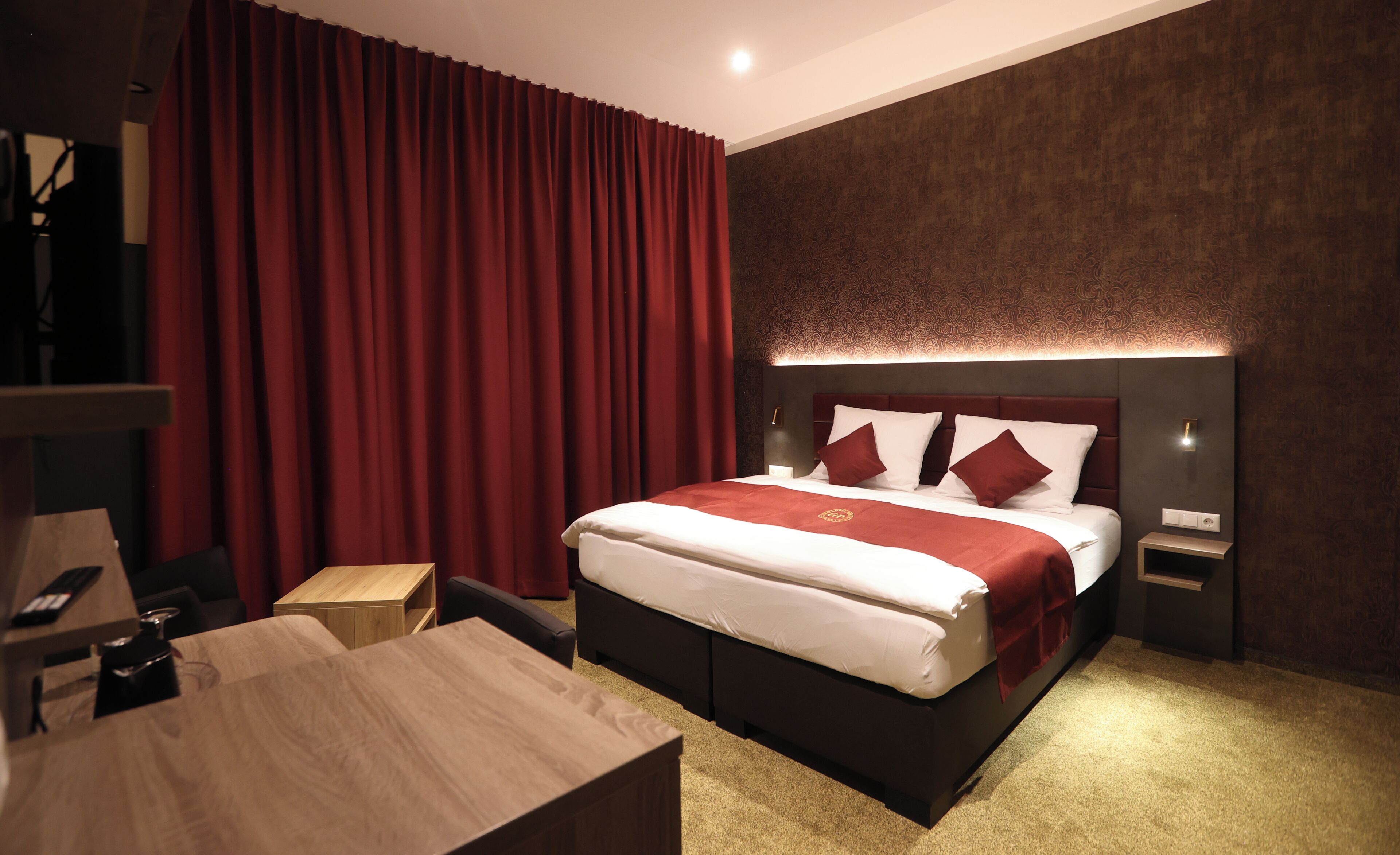 Ein modernes Hotelzimmer mit einem großen Bett, roten Vorhängen, einem Holztisch und einem Nachttisch mit Lampe.