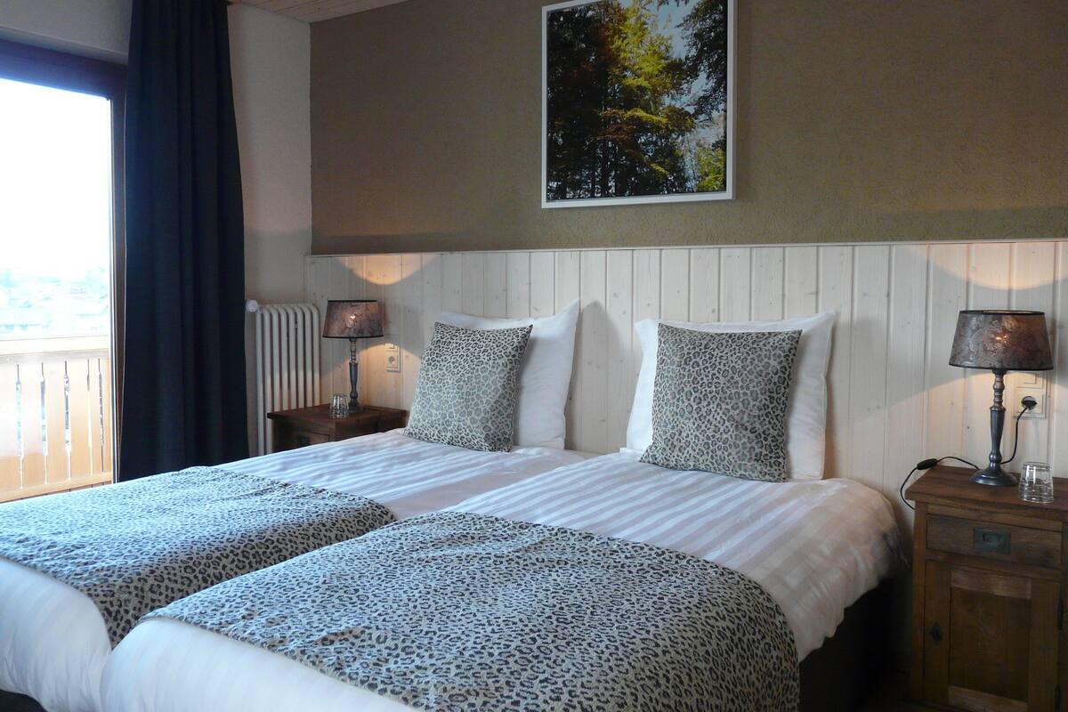 A hotel room with a large bed, two nightstands, a window with curtains, a radiator, and a painting on the wall.