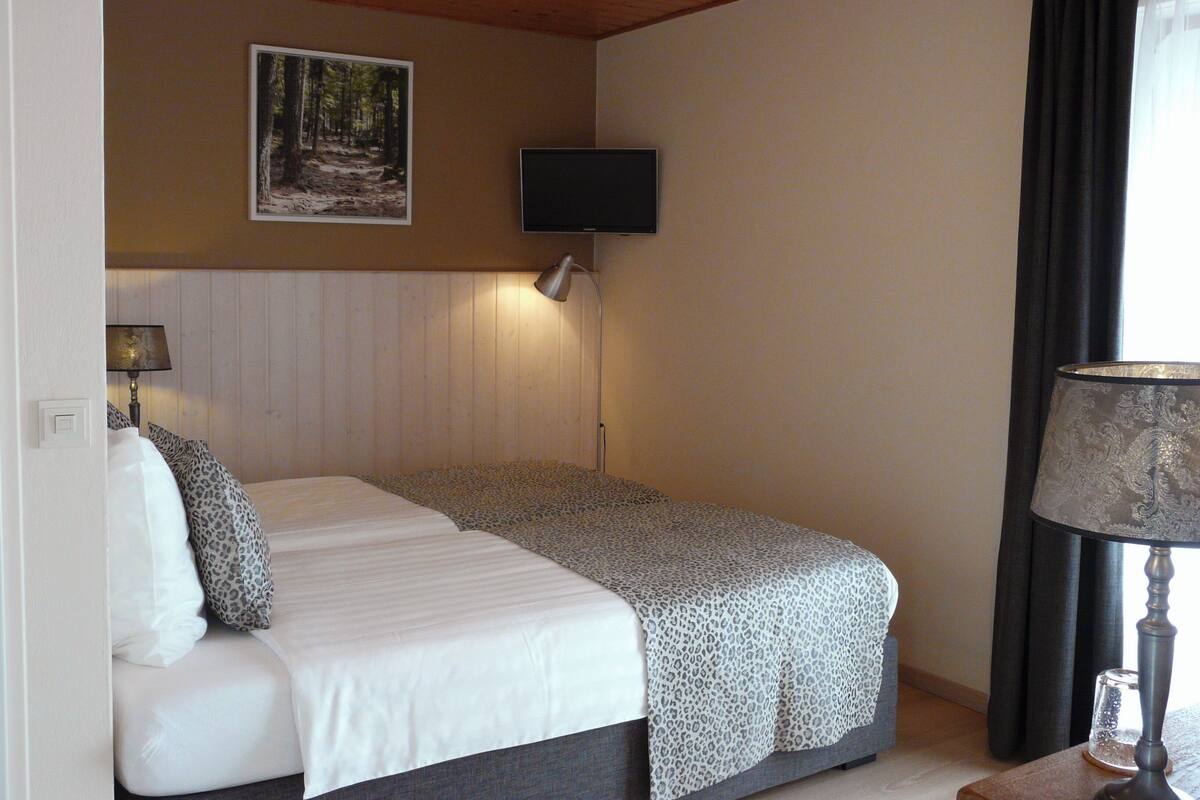 A bedroom with a bed, a TV mounted on the wall, a window with curtains, and a lamp on a bedside table.