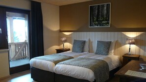 Ein Hotelzimmer mit einem großen Bett, zwei Nachttischen mit Lampen, einem Holz-Kopfteil, einem Bild von einem Wald und einem Balkon mit Geländer.
