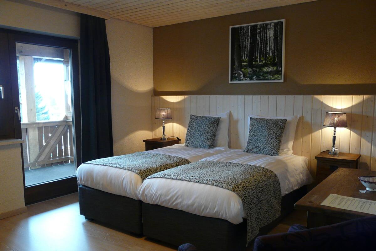 A hotel room with a large bed, two bedside tables with lamps, a wooden headboard, a painting of a forest, and a balcony with a railing.