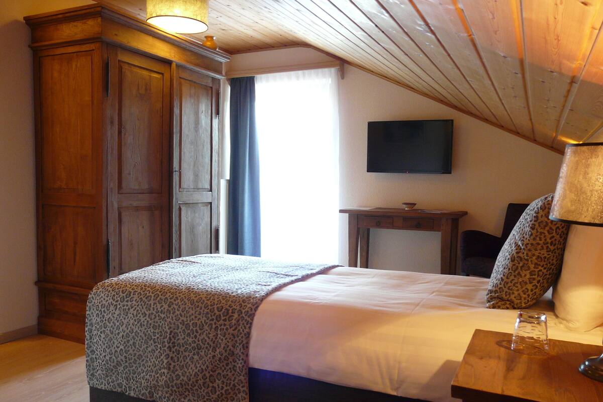 A bedroom with a bed, wooden wardrobe, a television on the wall, and a small table with a lamp.