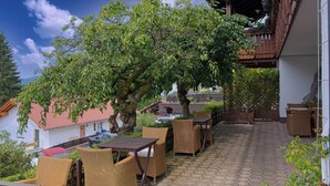 Eine Terrasse im Freien mit Korbmöbeln, einem Baum, der Schatten spendet, und Blick auf die umliegenden Gebäude.