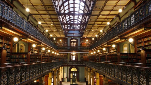 A grand library with a high ceiling, ornate railings, and rows of bookshelves.