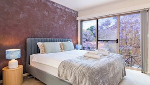 A bedroom with a large bed, a bedside lamp, and a view of trees through a sliding glass door.