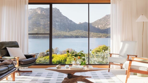 A room with a large window offering a view of a lake and mountains. There is a black leather chair, a wooden table, and a striped armchair. - Perfectly positioned! (Glamorgan-Spring Bay)