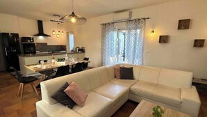 Ein modernes Wohnzimmer mit einer Eckcouch, einem Essbereich mit Tisch und Stühlen, einer Küche mit Geräten und einem großen Fenster mit Vorhängen.