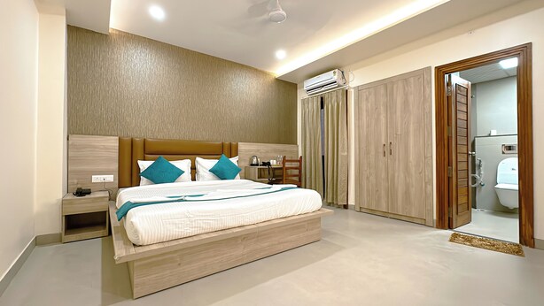A modern bedroom with a wooden headboard, a bed with white bedding and teal pillows, a bedside table, a nightstand, an air conditioning unit, a wooden wardrobe, and a bathroom visible through an open door. - Hotel Ananda Kashi (Varanasi)