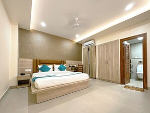 A modern bedroom with a wooden headboard, a bed with white bedding and teal pillows, a bedside table, a nightstand, an air conditioning unit, a wooden wardrobe, and a bathroom visible through an open door. - Hotel Ananda Kashi (Varanasi)