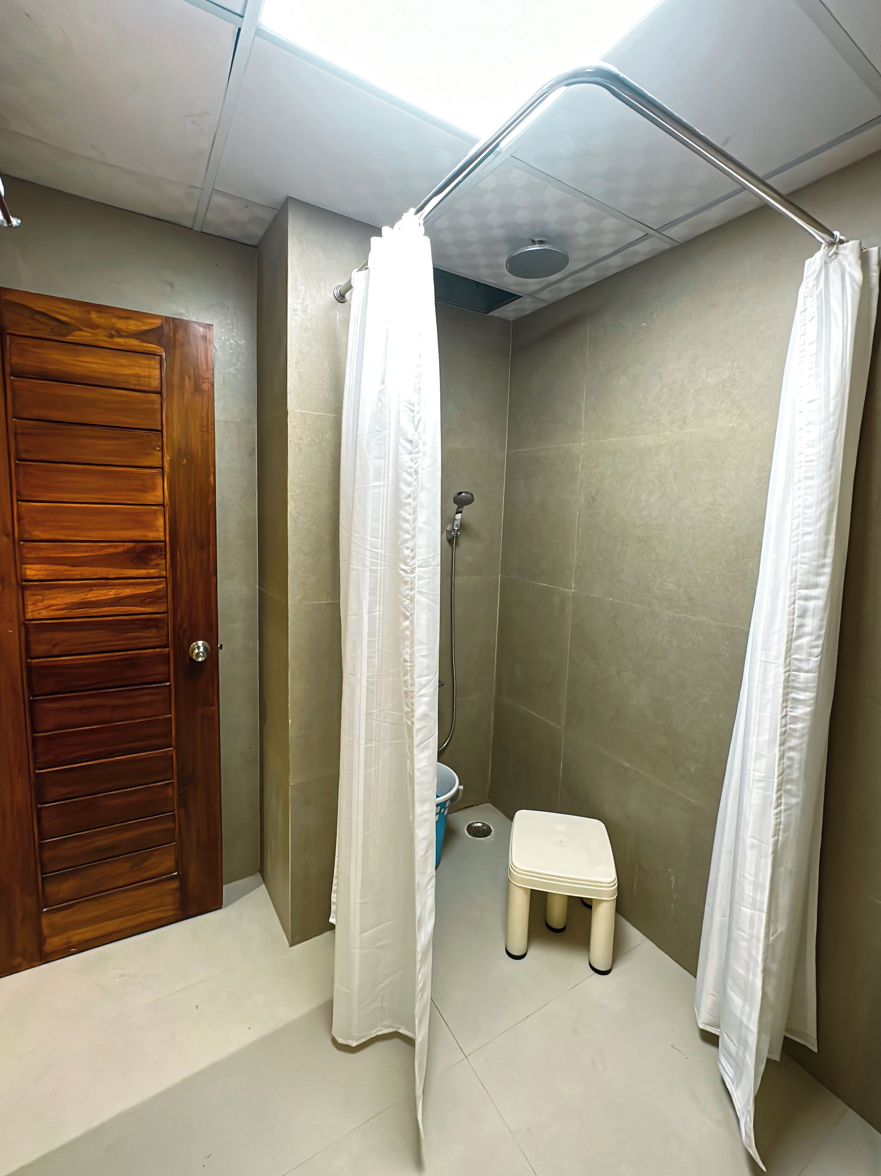Un espacio de ducha pequeño y cerrado con una puerta de madera, una cortina de ducha blanca y un taburete pequeño.