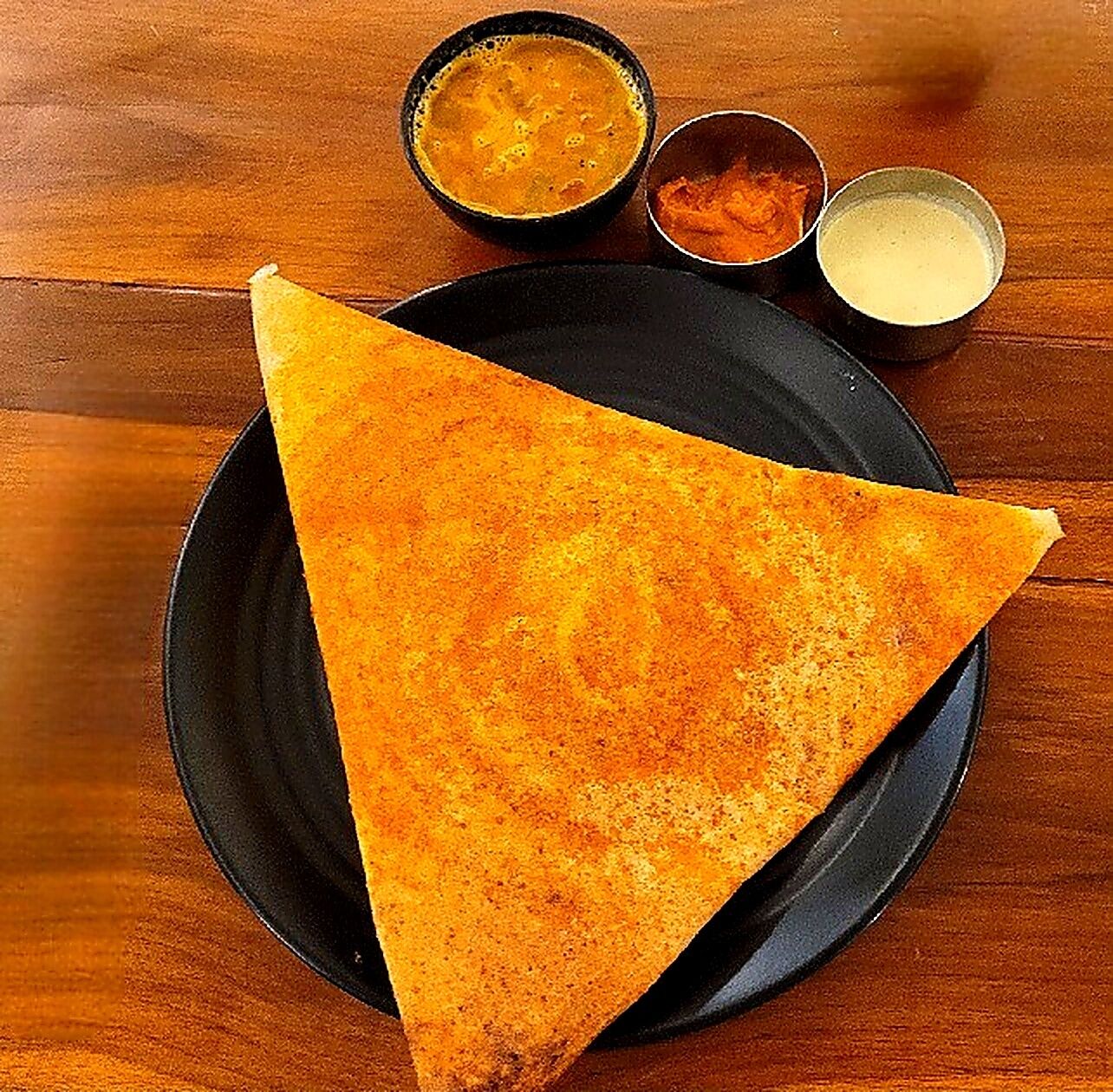 A triangular piece of food on a black plate, accompanied by two small bowls of sauces.