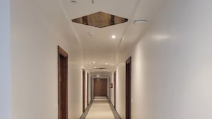 A long, narrow hallway with a light-colored carpet and dark-tiled floor. The walls are plain white, and there are several doors on either side. - Hotel Ananda Kashi (Varanasi)