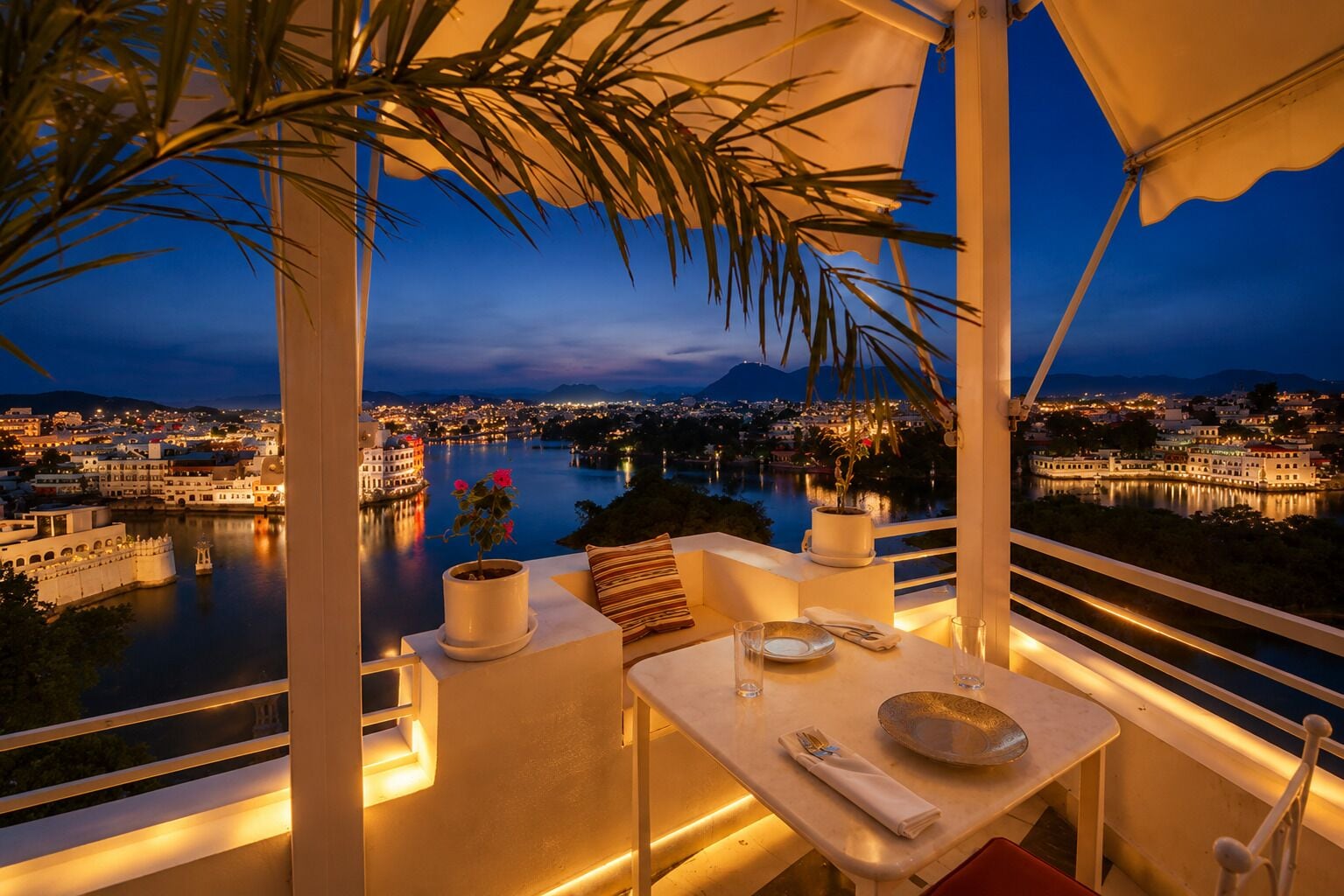 A dining area with a table set for two, overlooking a cityscape and a body of water at dusk.
