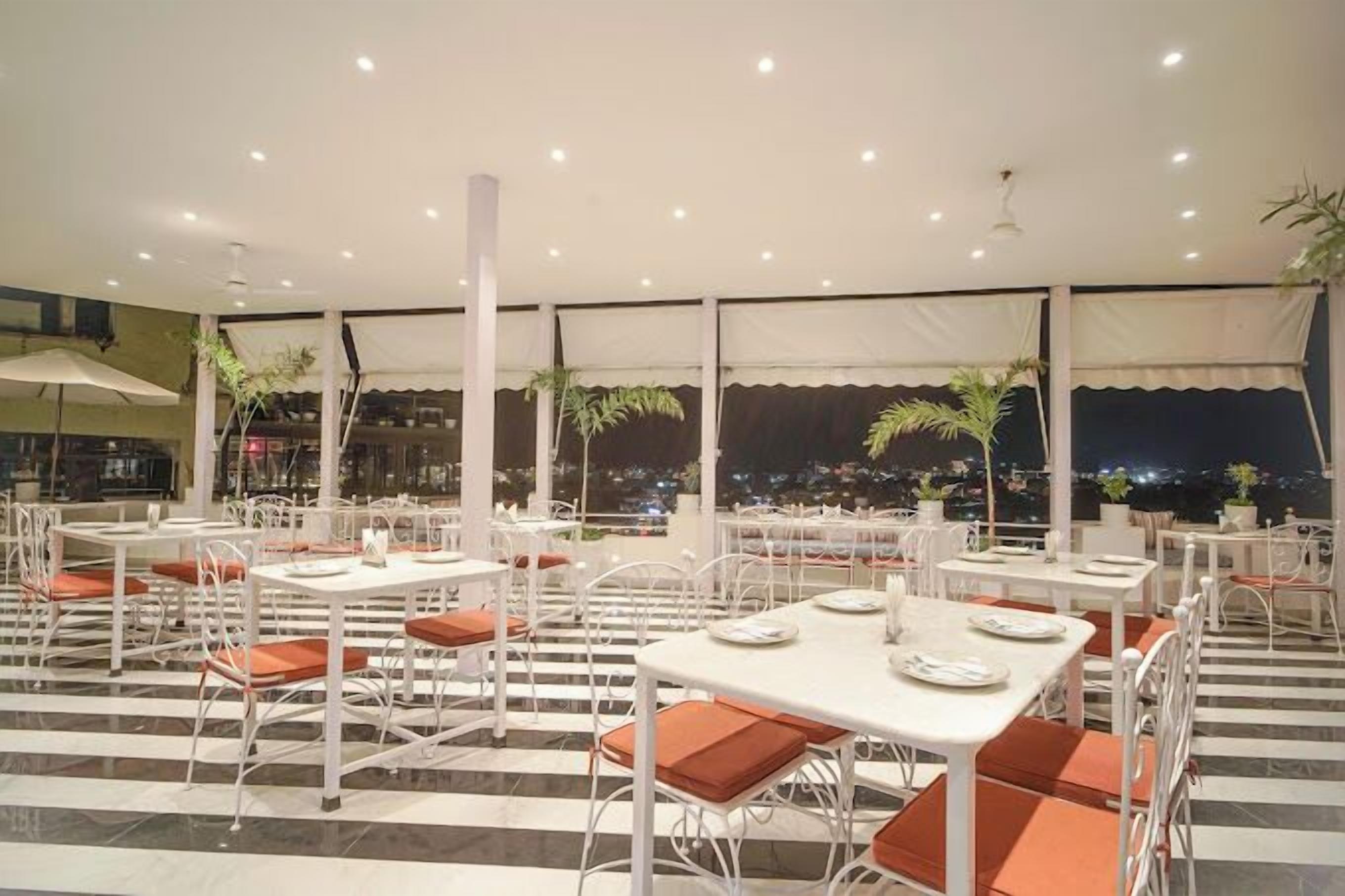 Ein Restaurant auf dem Dach mit weißen Tischen und orangefarbenen Sitzkissen, großen Fenstern mit Vorhängen und Blick auf die Stadt bei Nacht.